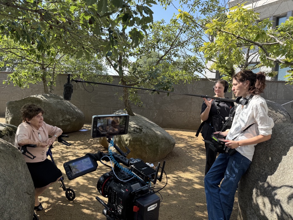Bronia sits with her walker among boulders and trees while the crew films, a boom overhead and Tillie leaning on a rock with a monitor.
