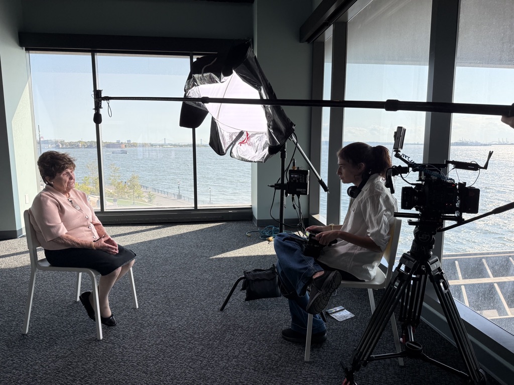Bronia seated for an interview by harbor windows, a crew member beside the camera and a softbox overhead.