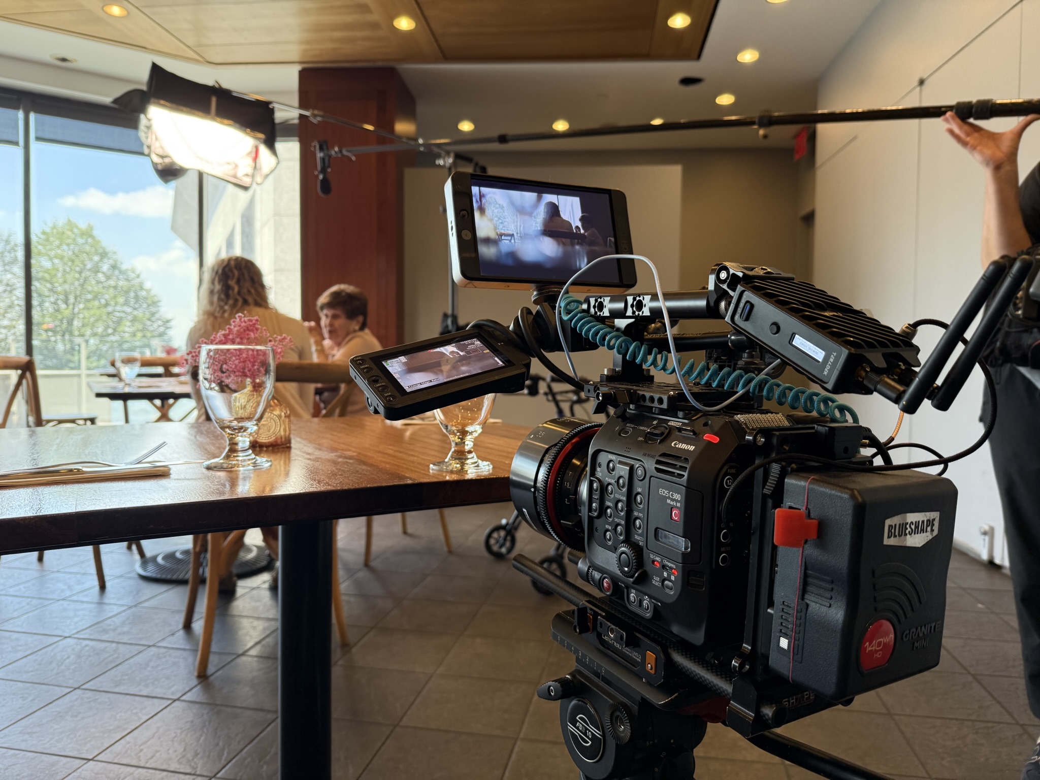 A Canon camera rig in the foreground with Bronia and a companion at a restaurant table behind it.