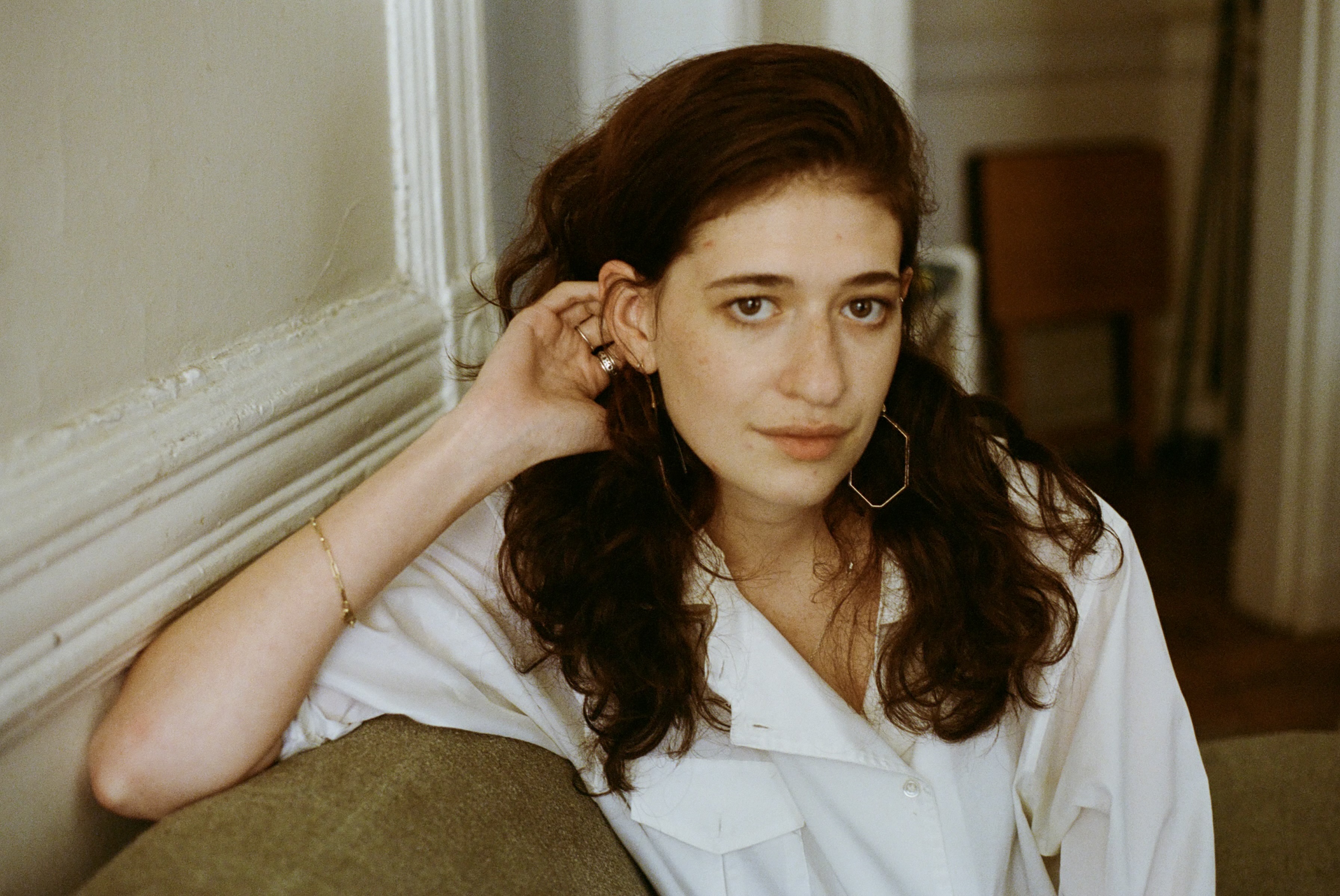Portrait of Tillie Simon in a white shirt, seated on a green sofa with one hand tucked behind her ear.