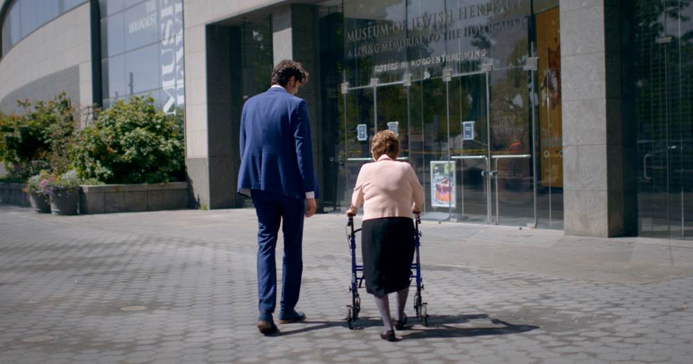 Isaac, in a blue suit, walks beside Bronia and her walker toward the entrance of the Museum of Jewish Heritage.