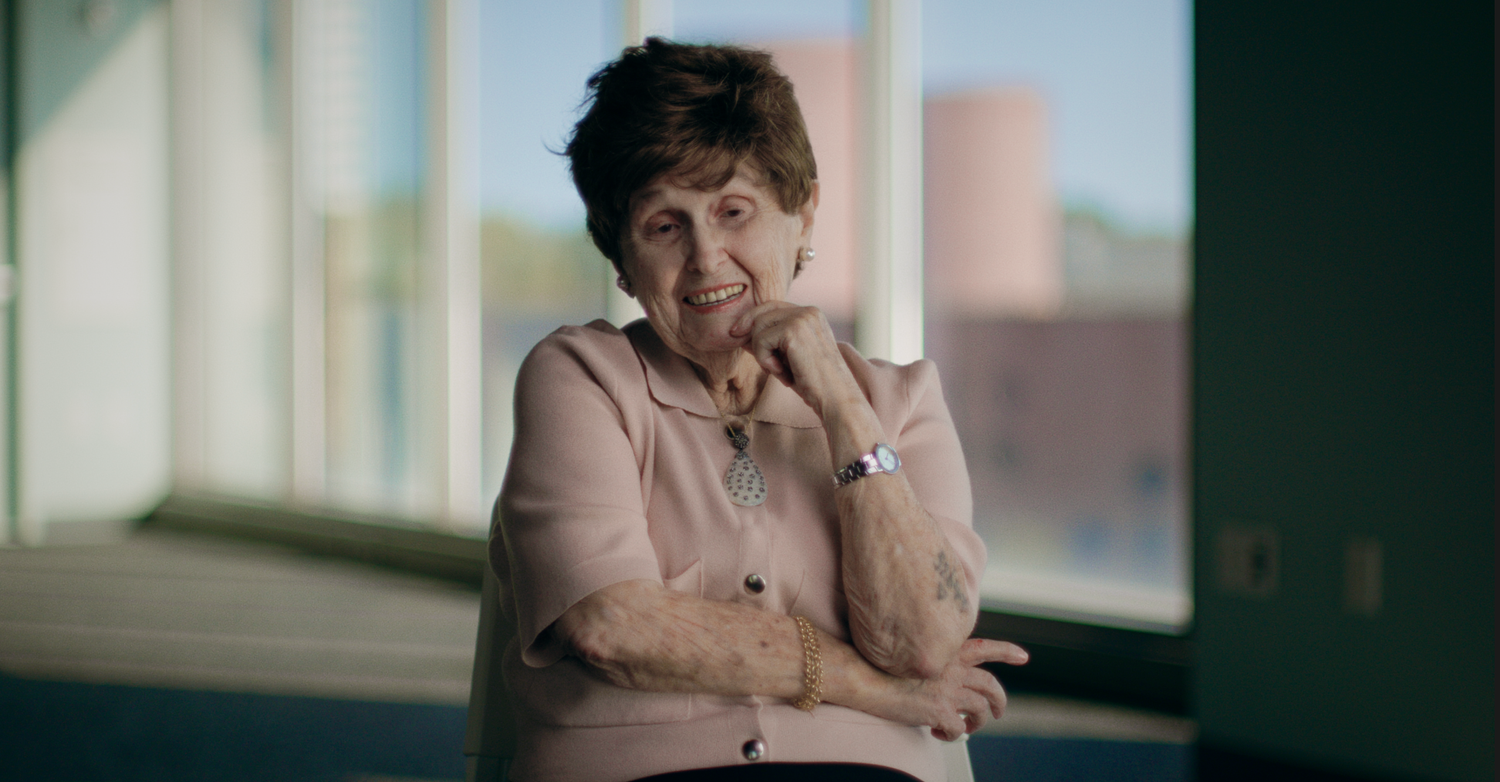 Bronia sits by a bright window, smiling, her hand at her chin and her arms folded.