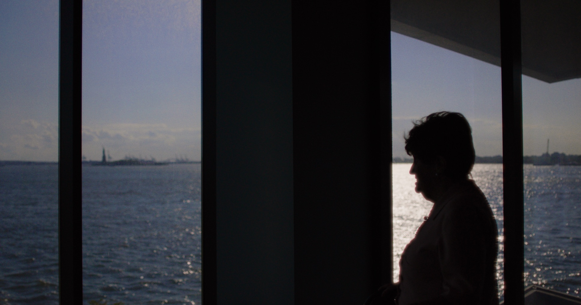 Bronia in silhouette by tall windows overlooking the harbor and the Statue of Liberty.